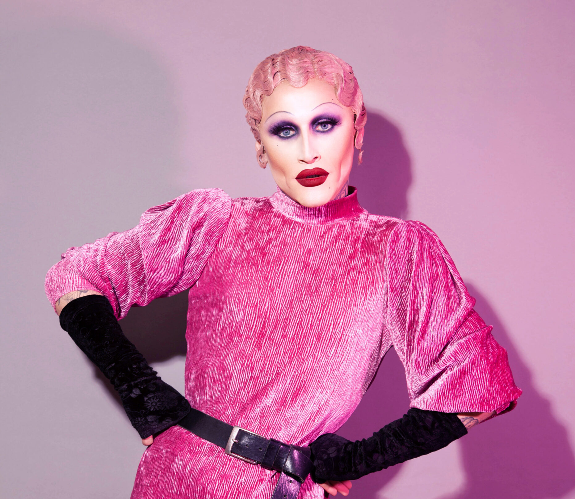 Mister Joe Black, pictured in this promotional photo wearing the infamous pink H&M dress from Ru Paul's Drag Race UK. Joe is also wearing black gloves and is stood against a photo backdrop.